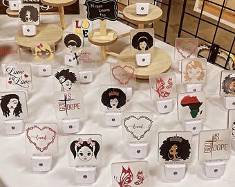 May include: A table display of various night lights featuring different designs. The designs include a butterfly, a heart, a crown, a cross, and a map of Africa. The night lights are all white and have a square shape. The text "God is dope" and "Love is Love" are visible on some of the night lights.