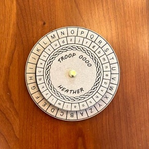 May include: A white circular cipher disk with two rotating rings. The outer ring has the letters of the alphabet, while the inner ring has the word "TROOP" and the numbers "0000". The inner ring also has the word "HEATHER" printed on it.