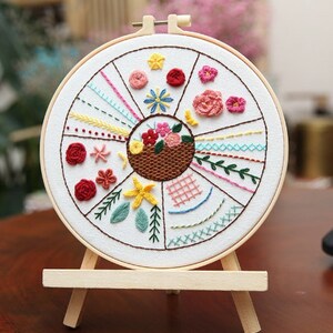 May include: A white embroidery hoop with a colorful floral design and various stitch patterns. The hoop is on a wooden easel.