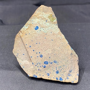 May include: A rough-hewn, irregularly shaped rock specimen. The stone has a tan base color with scattered, vibrant blue mineral deposits. Hints of green and blue-green hues are also visible. The rock is displayed against a dark background.