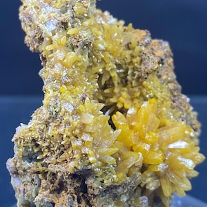 Op de afbeelding: Close-up van een mineraalmonster met clusters van heldergele, kristallijne structuren. De kristallen zijn gegroepeerd op een matrix van bruin en grijs gesteente. De algehele uitstraling is ruw en getextureerd, met een natuurlijke, aardse esthetiek.