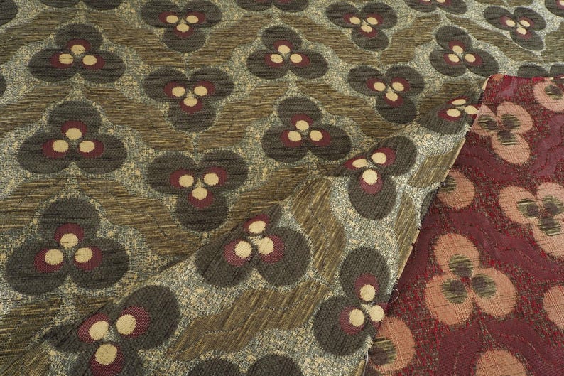 May include: Close-up of patterned fabric with a repeating floral design. The fabric features a textured background in shades of brown and gray, with dark gray floral shapes accented by burgundy and cream-colored details. A section of burgundy fabric is visible.