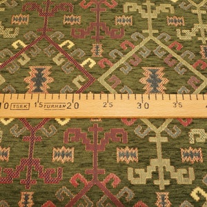 May include: Green fabric with a woven geometric pattern in shades of red, yellow, and blue. A wooden ruler with markings in inches and centimeters is placed across the fabric.