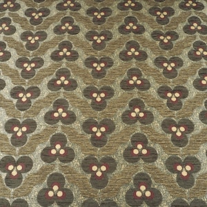 May include: Close-up of a patterned fabric with a repeating floral design. The pattern features dark gray, burgundy, and cream-colored floral shapes set against a textured, light brown background. The fabric has a woven appearance.