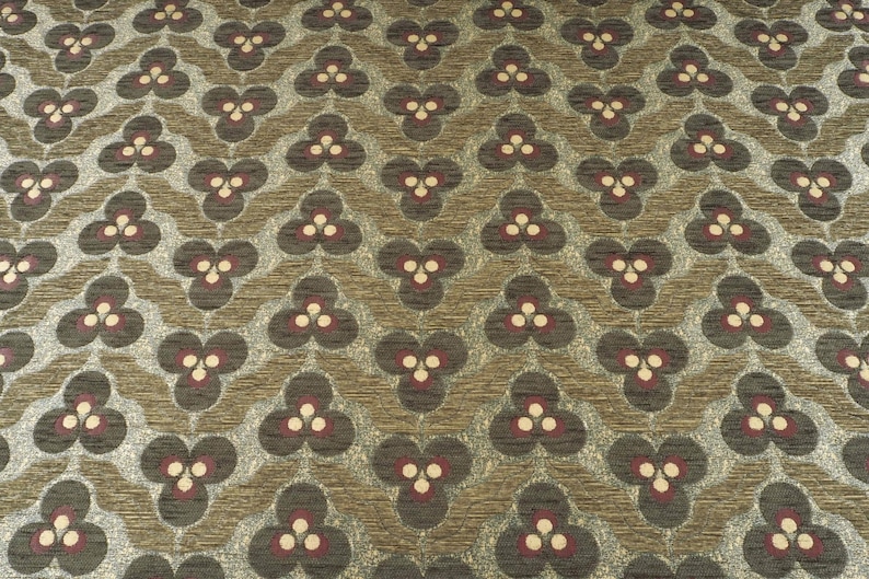 May include: Close-up of a patterned fabric with a repeating floral design. The pattern features dark gray, burgundy, and cream-colored floral shapes set against a textured, light brown background. The fabric has a woven appearance.