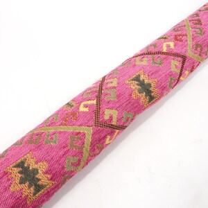 May include: Pink fabric with a geometric pattern in gold, brown, and black. The fabric is rolled up and has a textured surface.