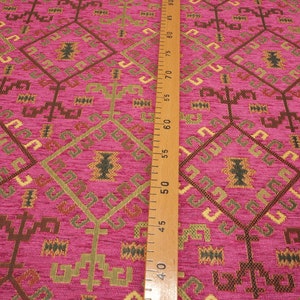 May include: Close-up of a pink fabric with a geometric pattern in shades of brown, gold, and green. The fabric is textured and has a woven look. A wooden ruler is visible on the right side of the image.