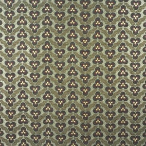 May include: A close-up of a patterned fabric with a repeating design. The pattern features dark brown clover-like shapes with three small yellow dots, set against a green background. The fabric has a textured appearance.