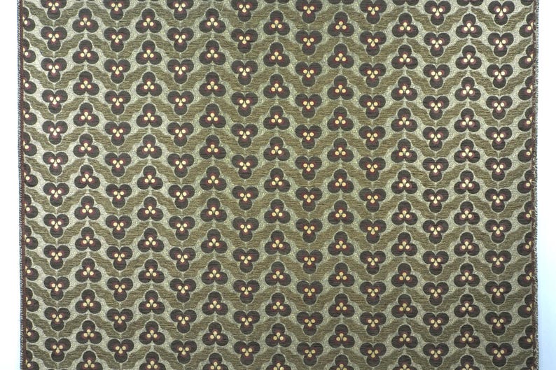 May include: A close-up of a patterned fabric with a repeating design. The pattern features dark brown clover-like shapes with three small yellow dots, set against a green background. The fabric has a textured appearance.