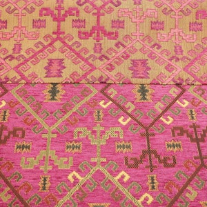 May include: A close-up of a pink and gold fabric with a repeating geometric pattern. The fabric has a textured surface and a woven look.