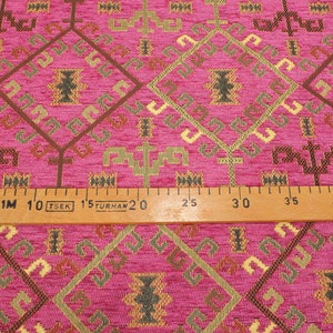 May include: Pink fabric with a geometric pattern in shades of brown, gold, and green. The fabric is measured with a ruler that reads "1M 10 TSEK 15 TURHAN 20 25 30 35 40 4".