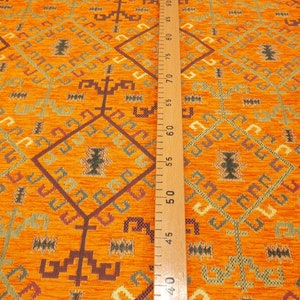 May include: Orange fabric with a geometric pattern in shades of brown, green, and cream. The fabric is measured with a wooden ruler.