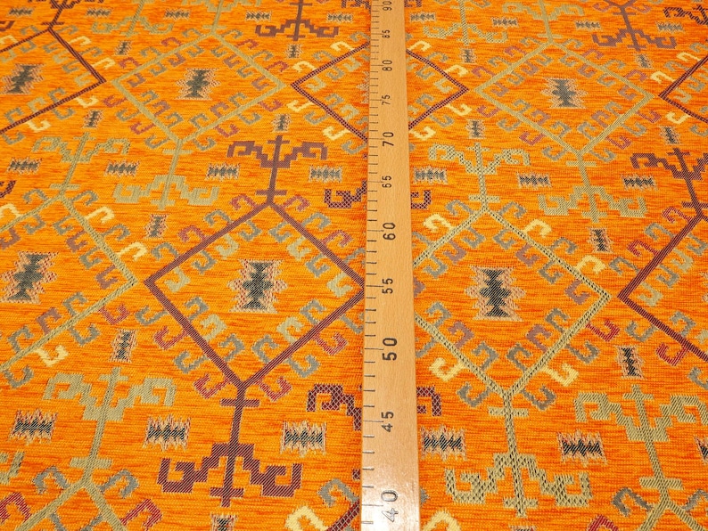 May include: Orange fabric with a geometric pattern in shades of brown, green, and cream. The fabric is measured with a wooden ruler.