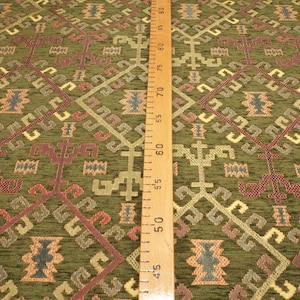 May include: Green fabric with a geometric pattern in shades of red, brown, gold, and gray. The fabric has a textured surface and is suitable for upholstery or crafting projects.