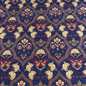 Puede incluir: Primer plano de un textil estampado con un diseño repetitivo. El patrón presenta formas florales y geométricas estilizadas en tonos de azul, marrón, rojo y crema. La textura general parece ser tejida, creando una superficie rica y detallada.