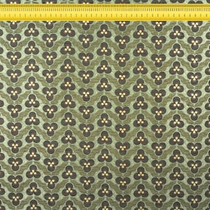 May include: Close-up of a fabric with a repeating floral pattern in shades of olive green and brown. The design features a stylized three-petal flower. A yellow ruler is placed along the edge of the fabric, showing inch measurements.