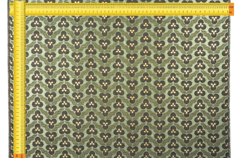 May include: Close-up of a fabric with a repeating floral pattern in shades of olive green and brown. The design features a stylized three-petal flower. A yellow ruler is placed along the edge of the fabric, showing inch measurements.