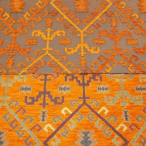 May include: Close-up of a patterned fabric with a geometric design in shades of orange, brown, and gray. The fabric has a textured surface and a woven appearance.
