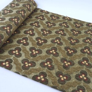 May include: Rolled fabric with a repeating floral pattern. The design features dark brown and cream-colored flowers on a textured olive green background. The fabric is likely used for upholstery or crafting.