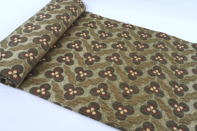 May include: Rolled fabric with a repeating floral pattern. The design features dark brown and cream-colored flowers on a textured olive green background. The fabric is likely used for upholstery or crafting.