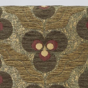 May include: Close-up of a decorative pillow with a repeating floral pattern. The design features dark brown, textured circles with red and cream-colored accents. The background has a woven, textured appearance in shades of brown and gold.