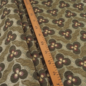 May include: Close-up of fabric with a repeating floral pattern. The flowers are dark gray with burgundy and cream-colored accents. The background is a textured olive green. A wooden ruler with inch markings is in the image.