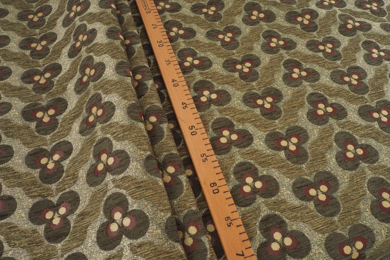 May include: Close-up of fabric with a repeating floral pattern. The flowers are dark gray with burgundy and cream-colored accents. The background is a textured olive green. A wooden ruler with inch markings is in the image.