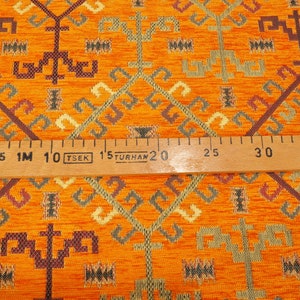 May include: Orange fabric with a geometric pattern in shades of brown, green, and cream. A wooden ruler with the text "1M TSEK 1.5 TURHAN" is placed on top of the fabric.