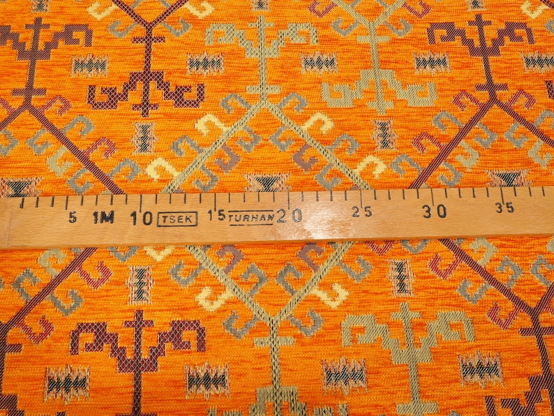 May include: Orange fabric with a geometric pattern in shades of brown, green, and cream. A wooden ruler with the text "1M TSEK 1.5 TURHAN" is placed on top of the fabric.
