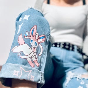 Puede incluir: Chaqueta vaquera azul claro con un gr&aacute;fico de dibujos animados de Sylveon en la manga. La chaqueta presenta un dise&ntilde;o desgastado con estrellas blancas y detalles de estrellas rosas. La manga est&aacute; doblada, revelando el borde sin rematar del denim.