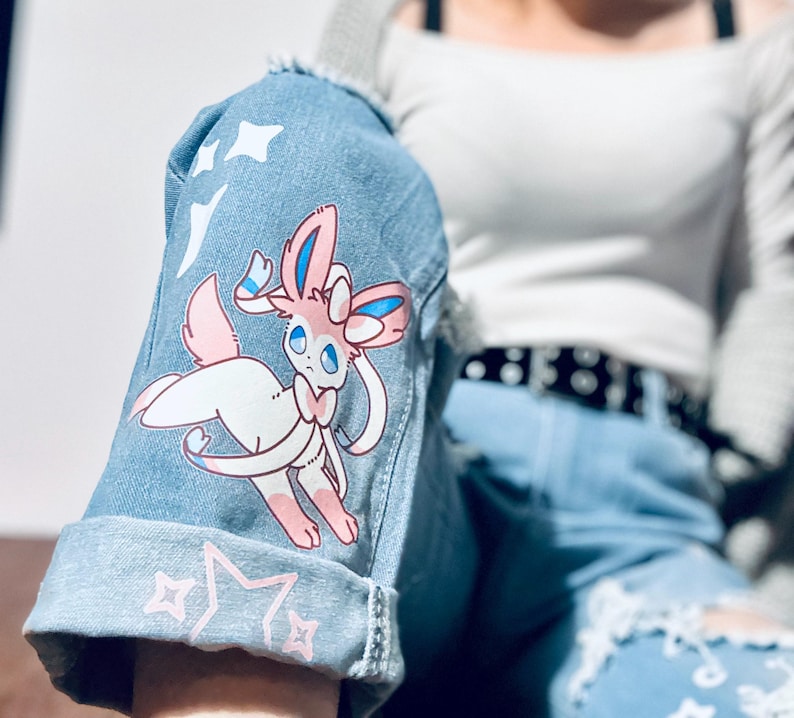 Puede incluir: Chaqueta vaquera azul claro con un gr&aacute;fico de dibujos animados de Sylveon en la manga. La chaqueta presenta un dise&ntilde;o desgastado con estrellas blancas y detalles de estrellas rosas. La manga est&aacute; doblada, revelando el borde sin rematar del denim.