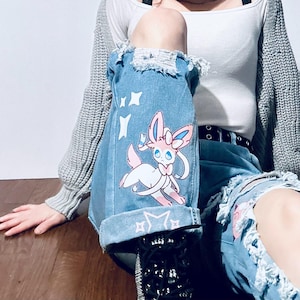 Puede incluir: Vaqueros de mezclilla azul claro con detalles desgastados y un gr&aacute;fico de dibujos animados de Sylveon. Los vaqueros tienen un dobladillo con dise&ntilde;o de estrellas y rodillas rasgadas. Sylveon es rosa, blanco y azul.