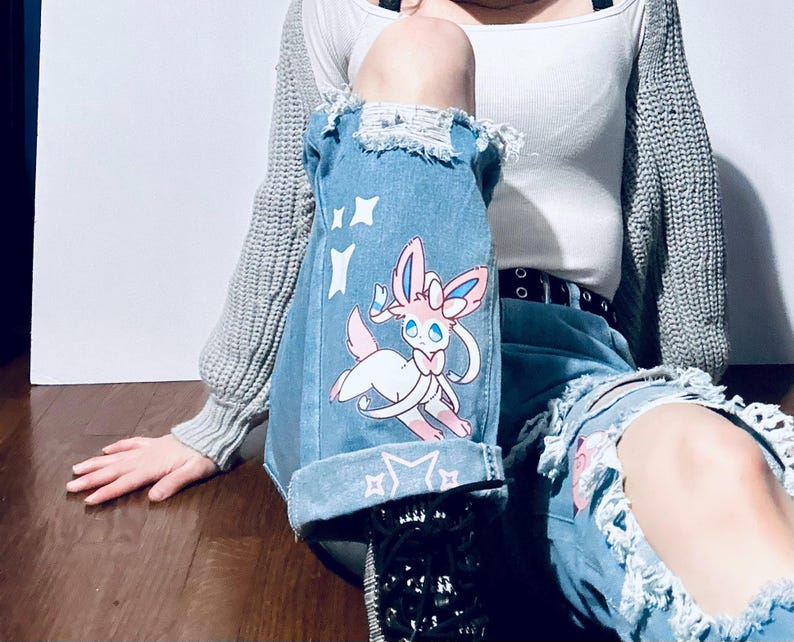 Puede incluir: Vaqueros de mezclilla azul claro con detalles desgastados y un gr&aacute;fico de dibujos animados de Sylveon. Los vaqueros tienen un dobladillo con dise&ntilde;o de estrellas y rodillas rasgadas. Sylveon es rosa, blanco y azul.