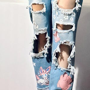 Puede incluir: Vaqueros azul claro con roturas y dise&ntilde;os de personajes de dibujos animados coloridos. Los vaqueros tienen m&uacute;ltiples roturas. Se combinan con botas de combate negras.