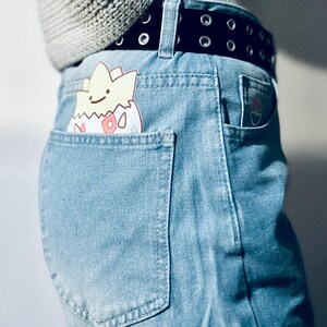 Puede incluir: Vaqueros de mezclilla azul claro con cintur&oacute;n negro y un personaje de dibujos animados asomando por el bolsillo trasero. El personaje es amarillo, blanco y rosa. Un peque&ntilde;o gr&aacute;fico est&aacute; en el bolsillo trasero derecho.