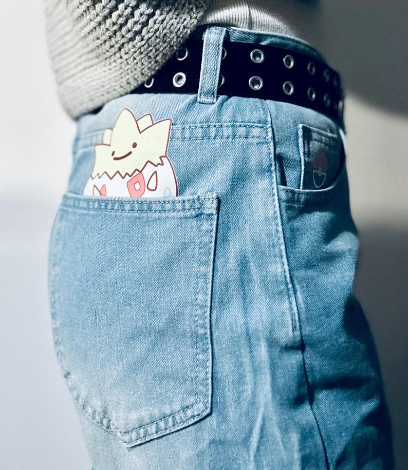 Puede incluir: Vaqueros de mezclilla azul claro con cintur&oacute;n negro y un personaje de dibujos animados asomando por el bolsillo trasero. El personaje es amarillo, blanco y rosa. Un peque&ntilde;o gr&aacute;fico est&aacute; en el bolsillo trasero derecho.
