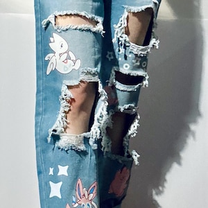 Puede incluir: Vaqueros azul claro desgastados con grandes rasgaduras y calcoman&iacute;as de personajes de dibujos animados. Los vaqueros tienen un cintur&oacute;n negro con ojales plateados y se combinan con botas a cuadros en blanco y negro.