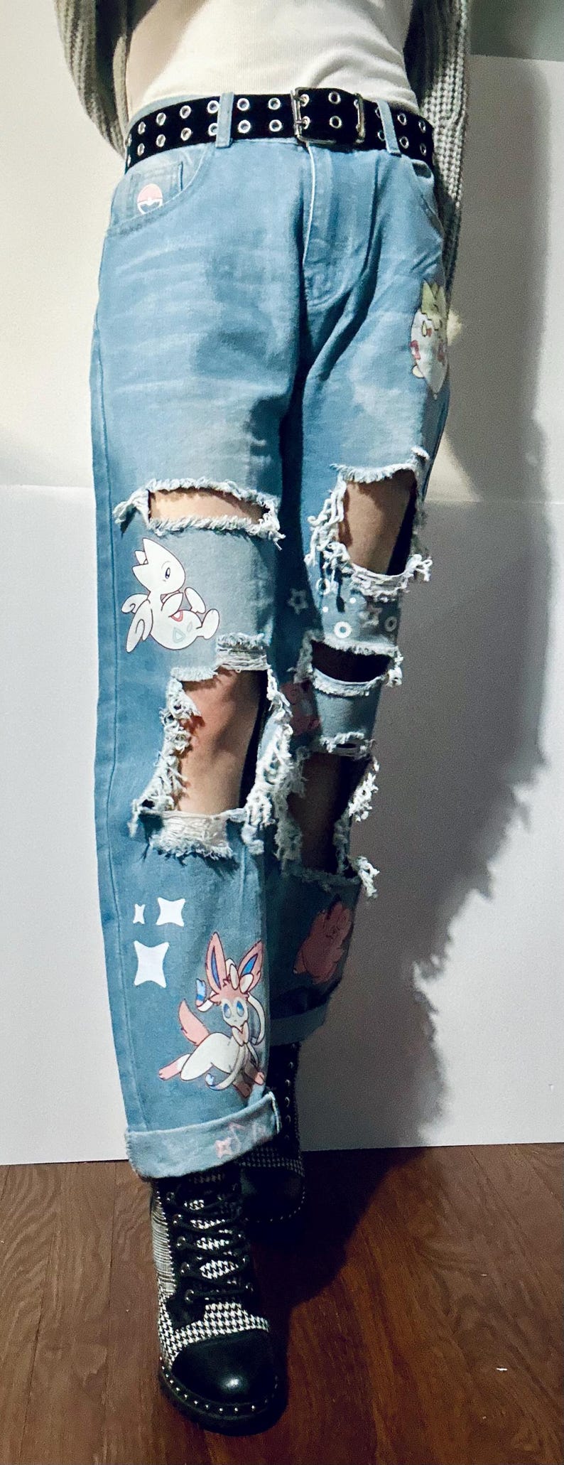 Puede incluir: Vaqueros azul claro desgastados con grandes rasgaduras y calcoman&iacute;as de personajes de dibujos animados. Los vaqueros tienen un cintur&oacute;n negro con ojales plateados y se combinan con botas a cuadros en blanco y negro.