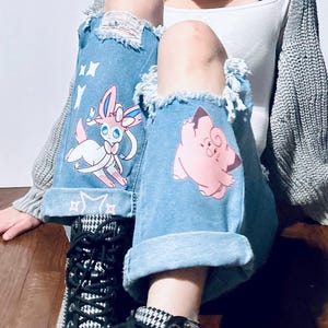 Puede incluir: Vaqueros de mezclilla azul claro con detalles desgastados y pu&ntilde;os doblados. Los vaqueros presentan gr&aacute;ficos de personajes de dibujos animados, incluyendo un Sylveon rosa y blanco y un Jigglypuff rosa. Los vaqueros se combinan con botas estampadas en blanco y negro.