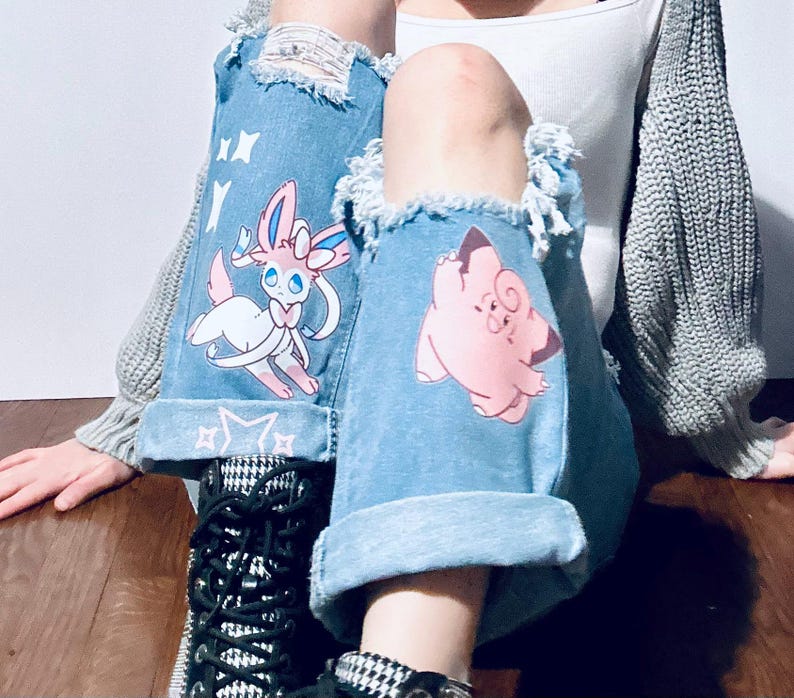 Puede incluir: Vaqueros de mezclilla azul claro con detalles desgastados y pu&ntilde;os doblados. Los vaqueros presentan gr&aacute;ficos de personajes de dibujos animados, incluyendo un Sylveon rosa y blanco y un Jigglypuff rosa. Los vaqueros se combinan con botas estampadas en blanco y negro.
