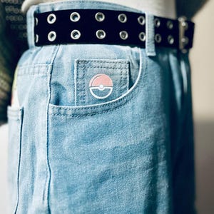 Puede incluir: Vaqueros azul claro con cintur&oacute;n negro con ojales. Un pin circular rosa y blanco est&aacute; sujeto al bolsillo delantero. El cintur&oacute;n tiene ojales plateados y una hebilla plateada.