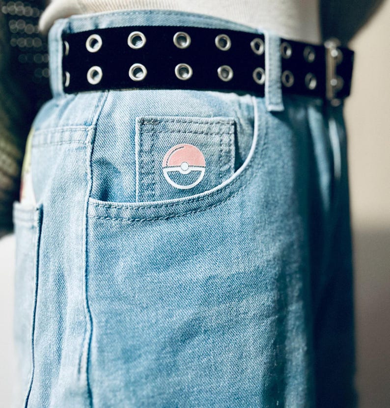 Puede incluir: Vaqueros azul claro con cintur&oacute;n negro con ojales. Un pin circular rosa y blanco est&aacute; sujeto al bolsillo delantero. El cintur&oacute;n tiene ojales plateados y una hebilla plateada.