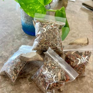 May include: Several small, clear plastic bags filled with dried herbs and spices. Each bag features a silver charm with a crescent moon and a star. The bags are arranged on a piece of driftwood, suggesting a natural or spiritual theme.