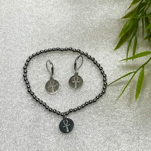 Op de afbeelding: Een zilveren armband met een zilveren bedel met een ankh-symbool. De armband is gemaakt van kleine zilveren kralen. Er zijn ook twee zilveren oorbellen met ankh-bedels.
