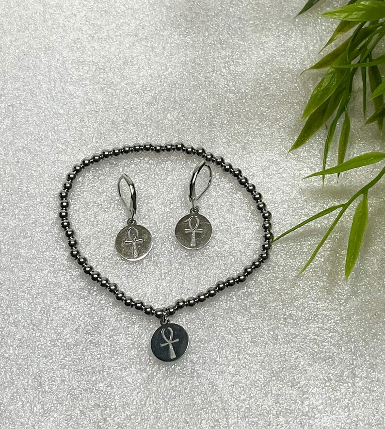 Op de afbeelding: Een zilveren armband met een zilveren bedel met een ankh-symbool. De armband is gemaakt van kleine zilveren kralen. Er zijn ook twee zilveren oorbellen met ankh-bedels.