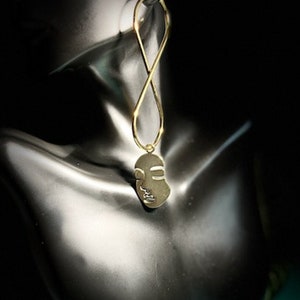 Op de afbeelding: Gouden, gezichtsvormige hanger-oorring met een gelust ontwerp. De oorring hangt aan het oor van een mannequin.