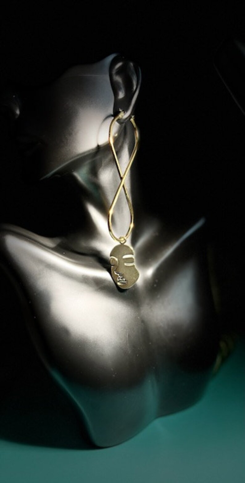 Op de afbeelding: Gouden, gezichtsvormige hanger-oorring met een gelust ontwerp. De oorring hangt aan het oor van een mannequin.
