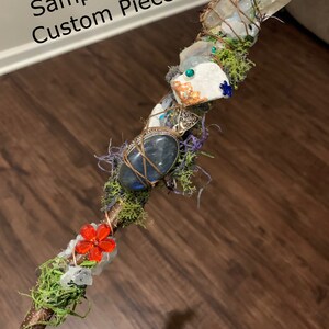 May include: A wooden wand with various crystals, moss, and a red flower charm. The wand is wrapped with copper wire and has a natural wood finish.
