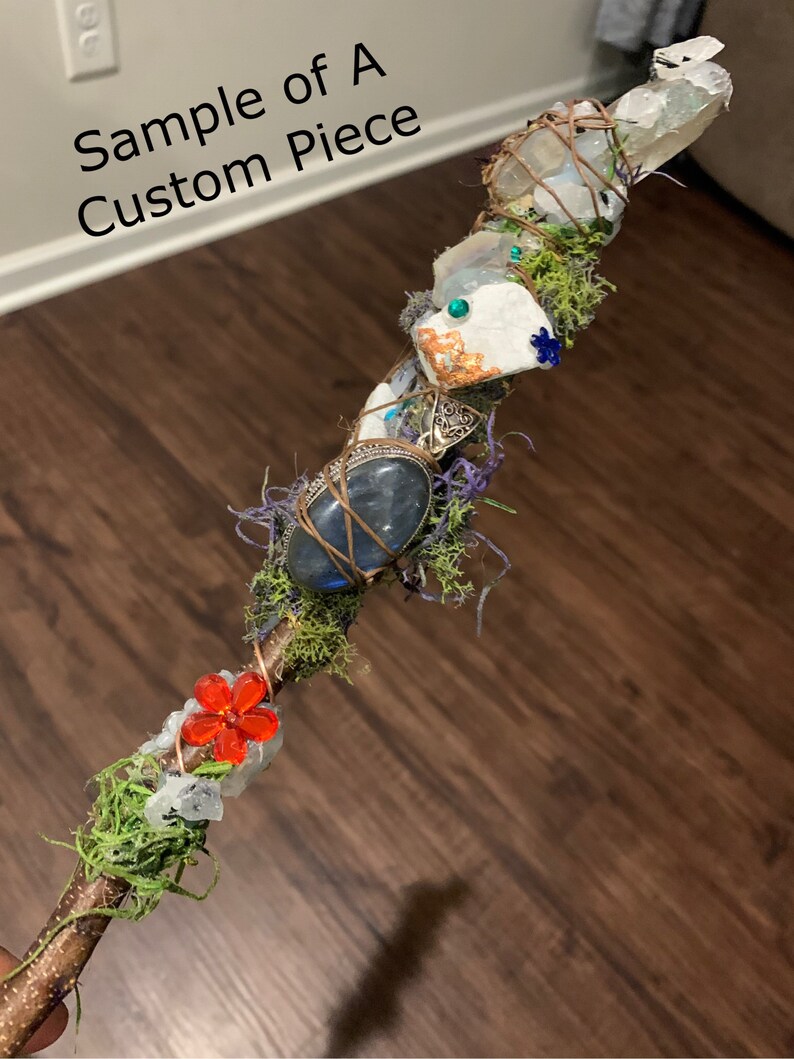 May include: A wooden wand with various crystals, moss, and a red flower charm. The wand is wrapped with copper wire and has a natural wood finish.