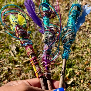 May include: Three wooden wands wrapped in colorful wire, feathers, and crystals. The wands are held in a hand with blue nail polish.